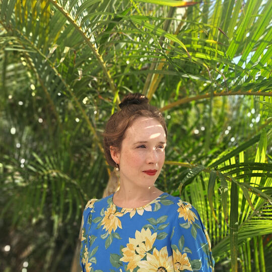 The image shows a white woman wearing a blew dress with sunflowers on posing in front of a palm tree.